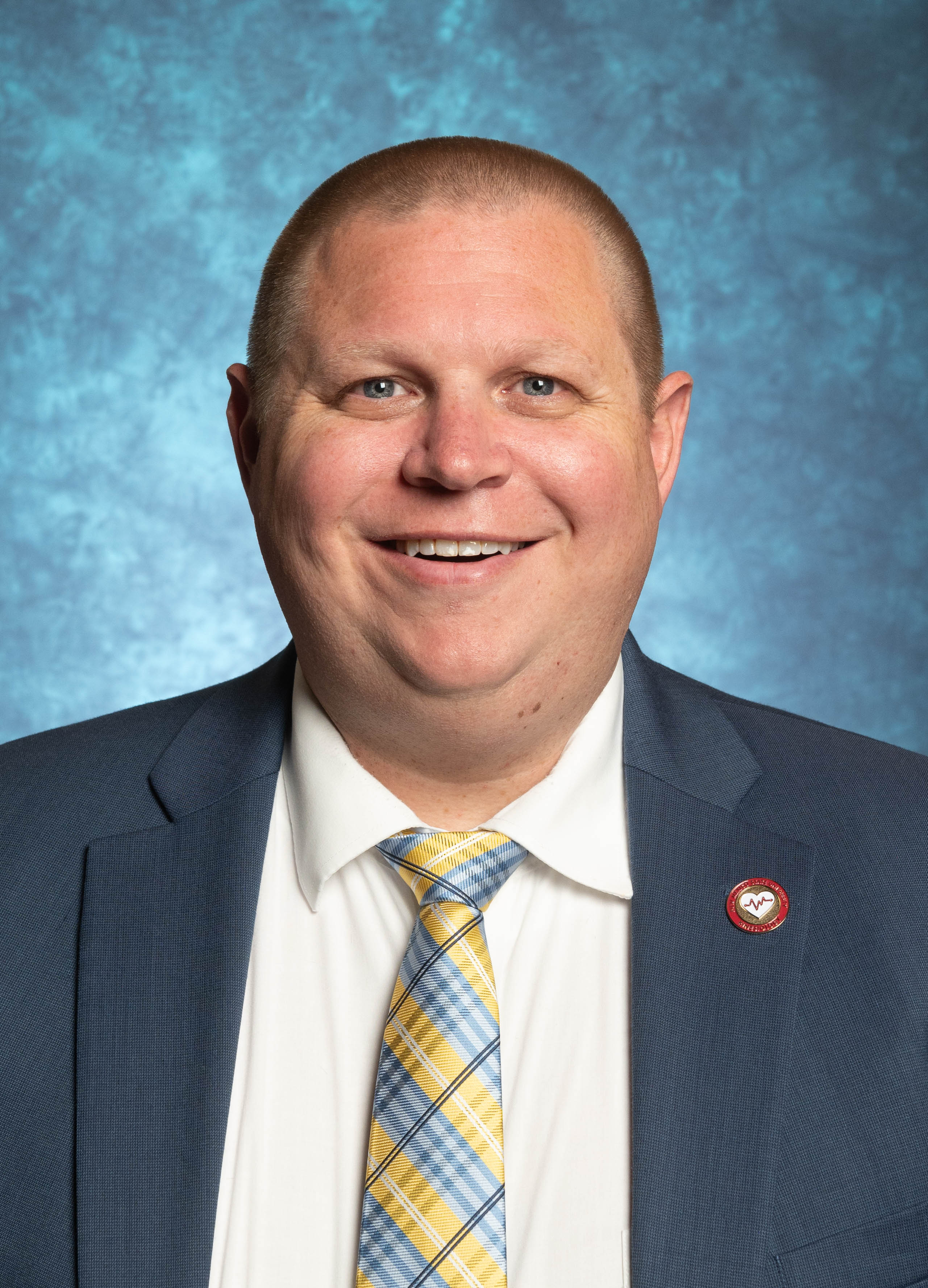 Head-and-shoulders portrait of a smiling man in a blue suit and patterned yellow-and-blue tie against a mottled blue background.